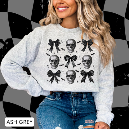 Woman wearing an ash grey crewneck sweatshirt featuring a coquette skull design with black bows, bats and gothic Halloween aesthetic.