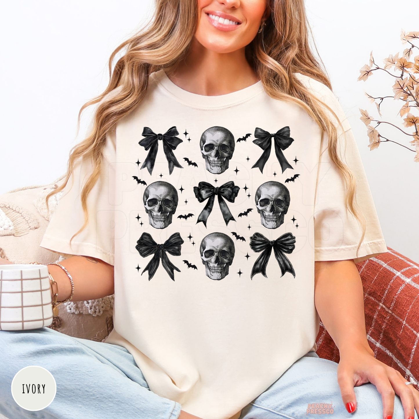 Woman wearing an ivory Comfort Colors t-shirt with a coquette skull graphic featuring black bows, bats and gothic Halloween artwork.