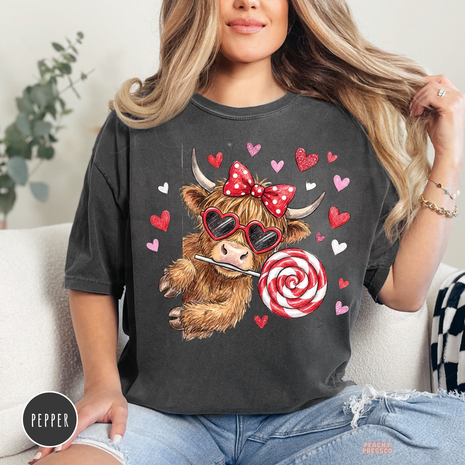 Cute coquette cow wearing heart-shaped sunglasses, a red polka dot bow, and holding a swirled lollipop on a pepper Comfort Colors Valentine's shirt.