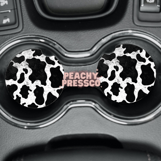 Black and white cow print ceramic car coasters displayed in vehicle cup holders.