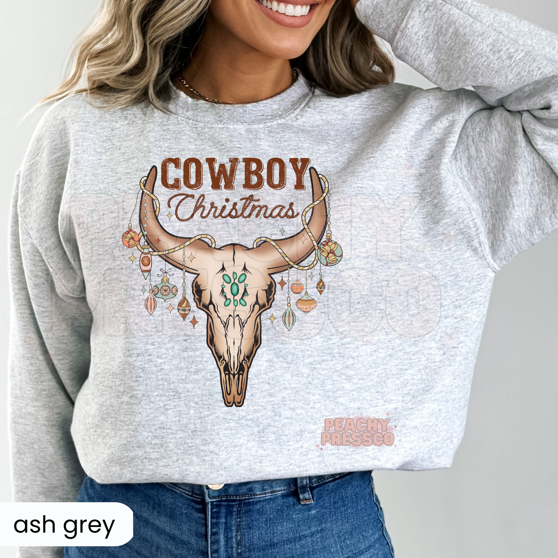 A woman wearing a ash gray sweatshirt with a 'Cowboy Christmas' graphic featuring a deer skull and ornaments.