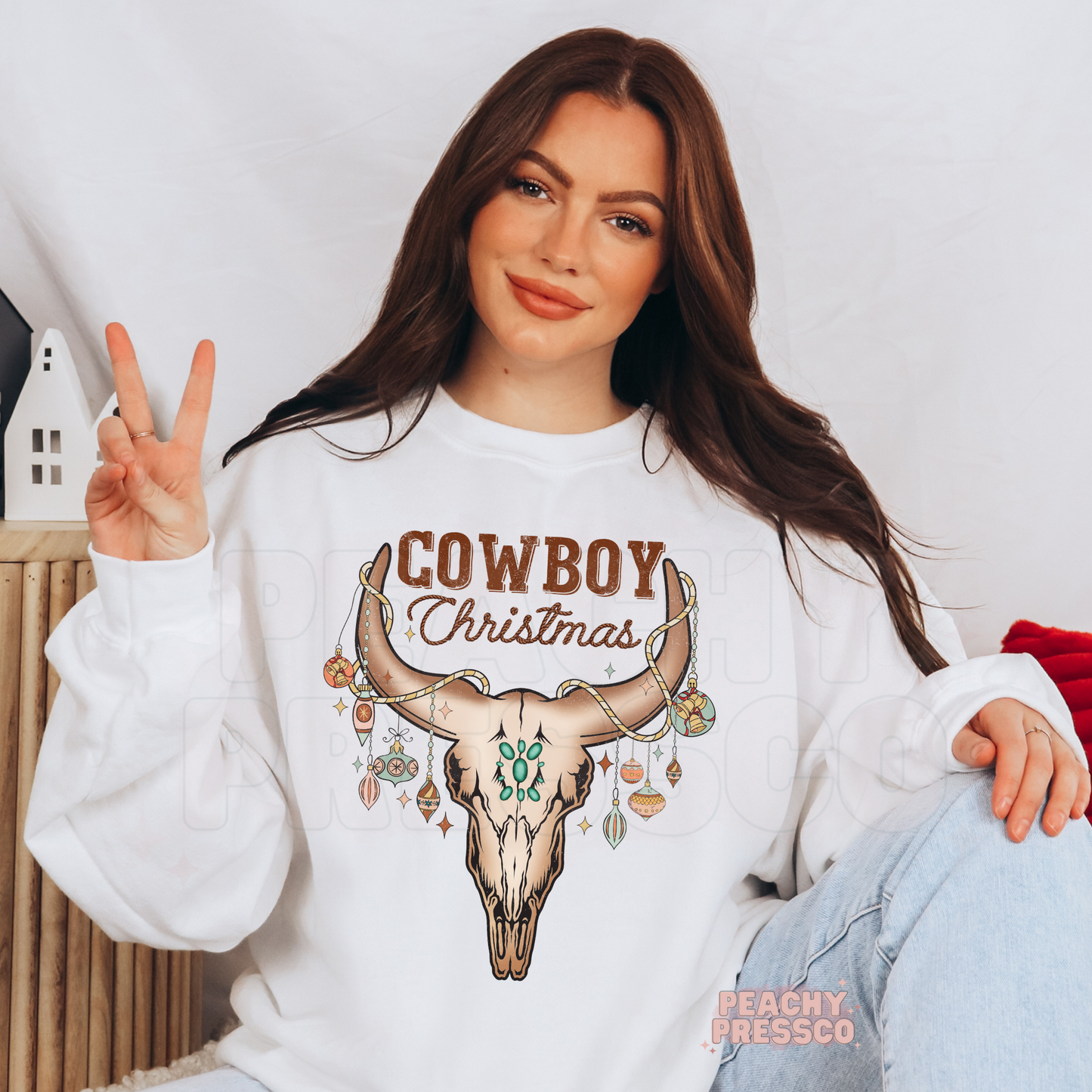 A woman wearing a white crewneck sweatshirt with a 'Cowboy Christmas' graphic featuring a decorated skull and ornaments, posing in a cozy indoor setting.
