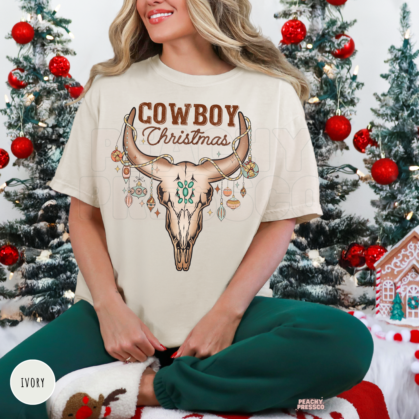 A woman wearing an ivory graphic t-shirt with a 'Cowboy Christmas' design featuring a skull and ornaments, posed in front of decorated Christmas trees.