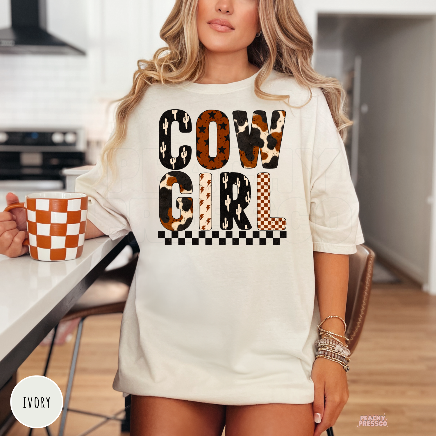 Ivory western t-shirt featuring a bold Cowgirl graphic with cow print, cactus, checkerboard, and lightning bolt designs for a country-inspired look.