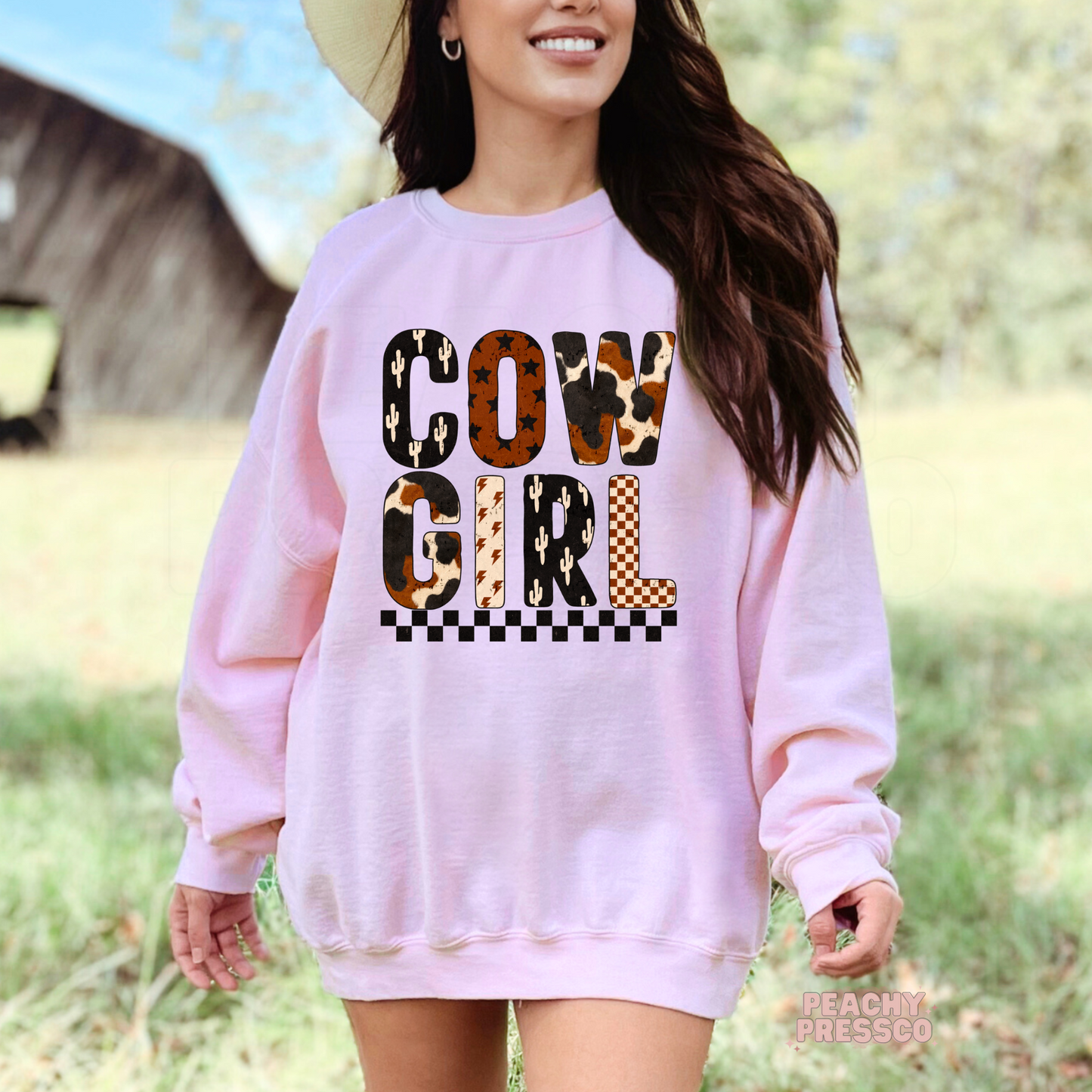 Woman wearing a light pink sweatshirt with a bold Cowgirl western graphic featuring cow print, cactus, checkerboard, and lightning bolt patterns.
