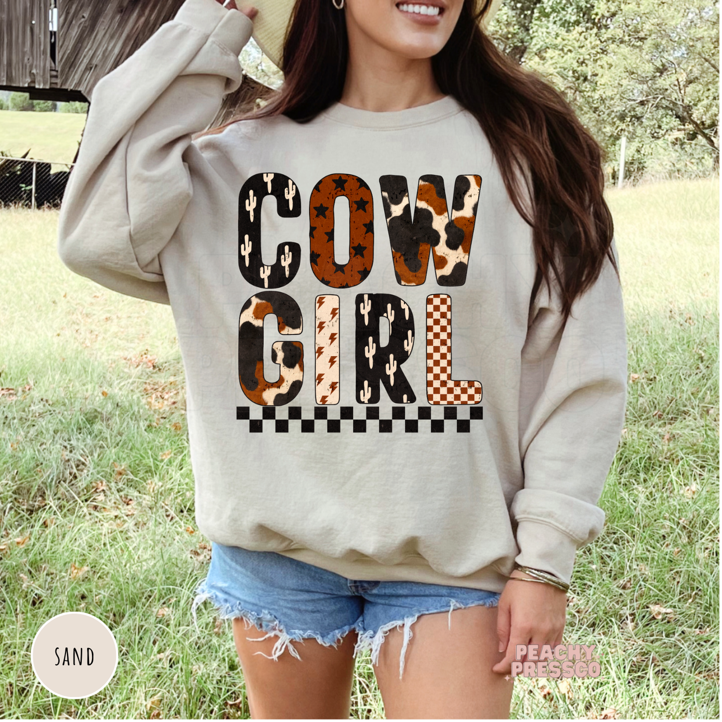 Sand colored western sweatshirt featuring a Cowgirl graphic with cow print, cactus, checkerboard, stars, and lightning bolt accents.