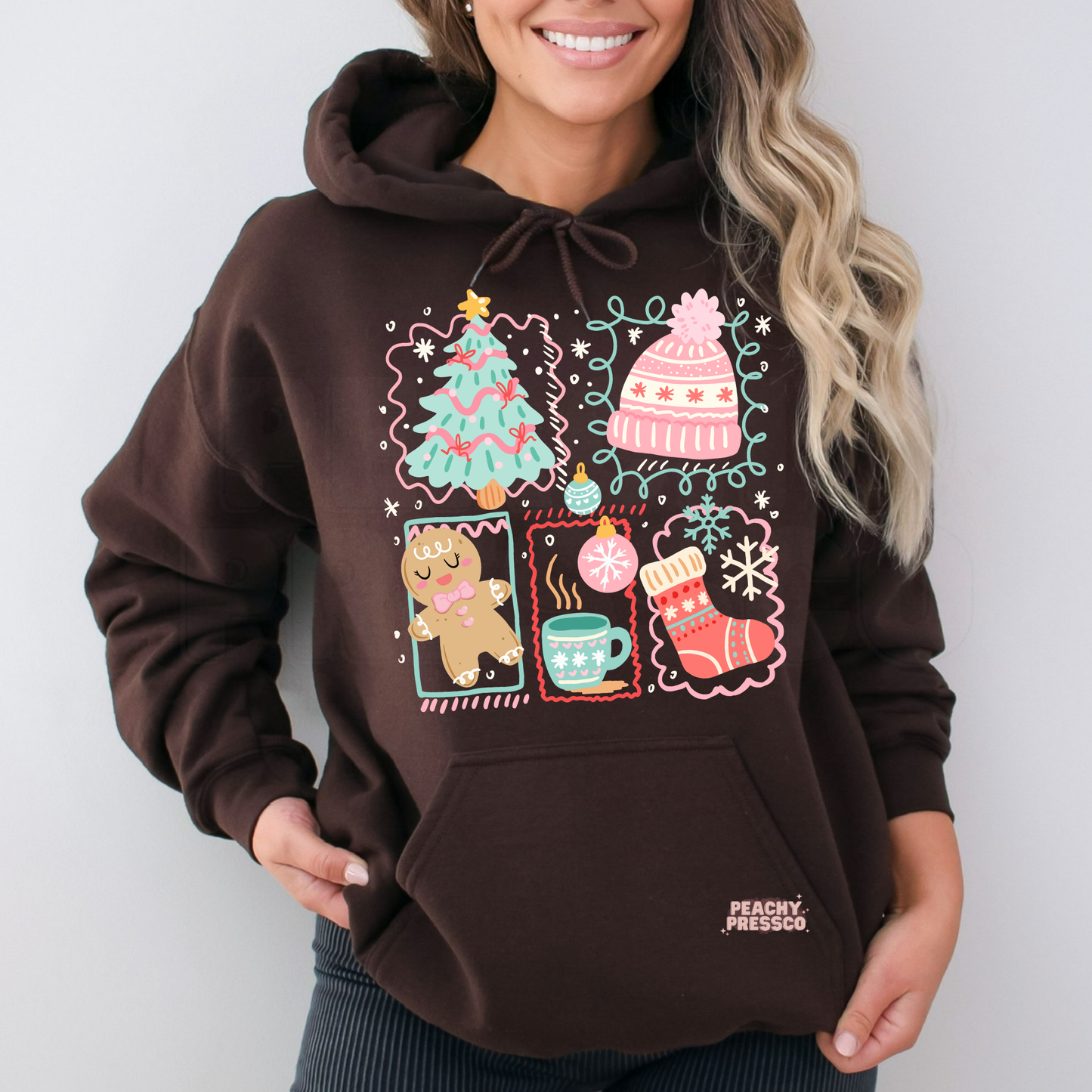A woman wearing a dark brown pullover hoodie with a colorful Christmas-themed graphic print on the front, featuring a tree, hat, gingerbread cookie, mug, and stocking. 