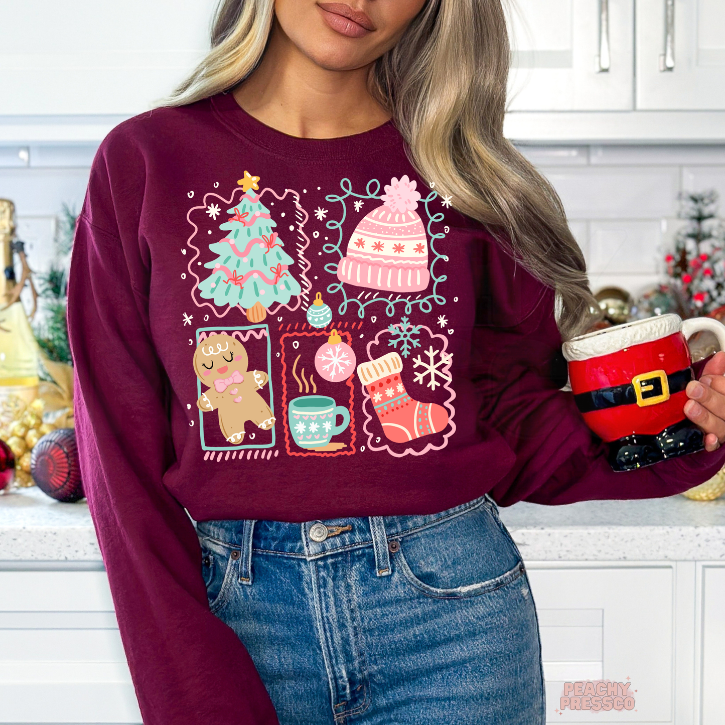 A woman wearing a maroon crewneck sweatshirt with a colorful Christmas-themed graphic print, paired with blue jeans and holding a Santa-themed mug in a kitchen setting.