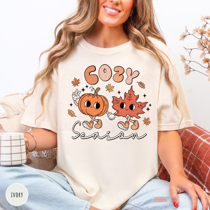 Woman wearing an ivory Comfort Colors t-shirt featuring a playful Cozy Season pumpkin and leaf graphic in a cute retro autumn style.