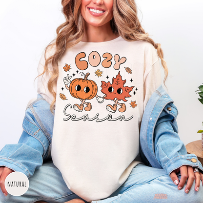 Woman wearing a natural shirt with a whimsical Cozy Season design featuring adorable pumpkin and leaf characters surrounded by autumn leaves.