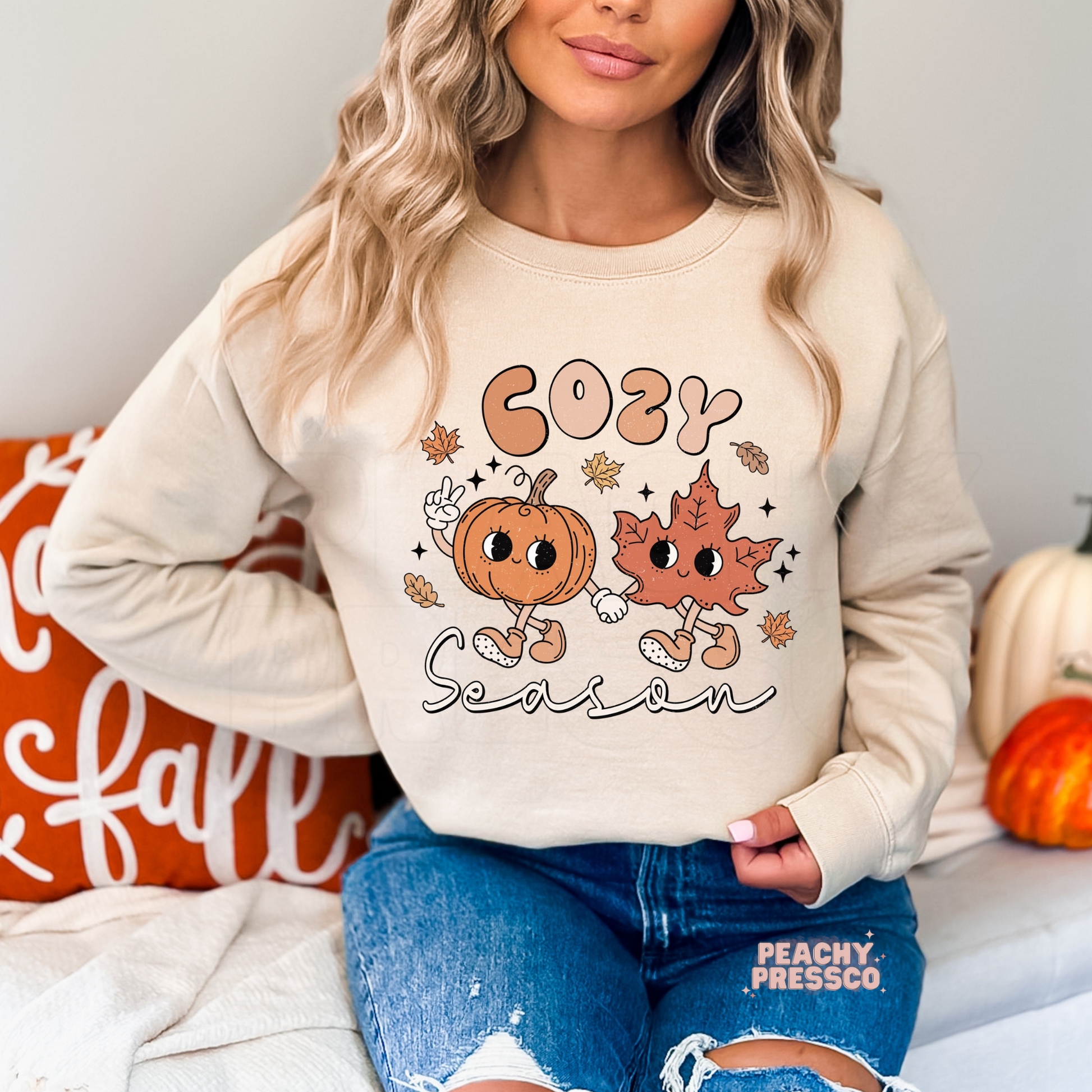 Woman wearing a sand crewneck sweatshirt with a Cozy Season fall graphic featuring smiling pumpkin and leaf characters holding hands.