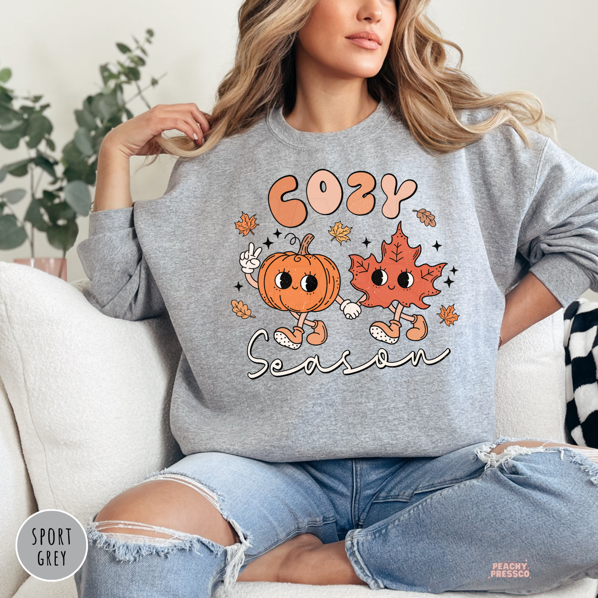 Woman wearing a sport grey crewneck sweatshirt featuring a cute "Cozy Season" design with a smiling pumpkin and leaf holding hands.