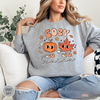 Woman wearing a sport grey crewneck sweatshirt featuring a cute "Cozy Season" design with a smiling pumpkin and leaf holding hands.
