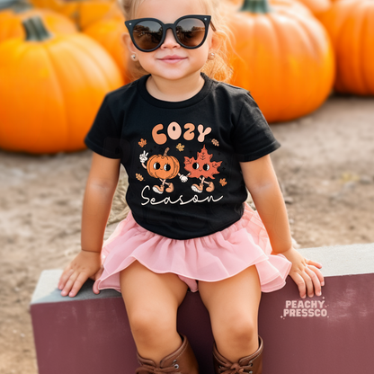 Toddler girl wearing a black "Cozy Season" fall t-shirt with cute pumpkin and maple leaf characters while sitting at a pumpkin patch.