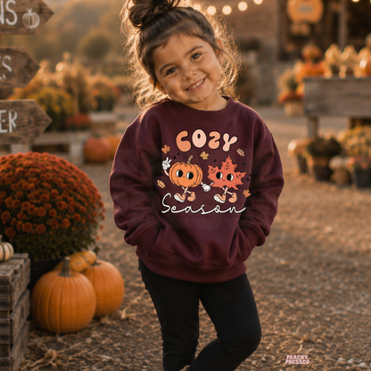 Cute toddler girl in a maroon "Cozy Season" sweatshirt featuring pumpkin and maple leaf characters during a cozy autumn pumpkin patch visit.