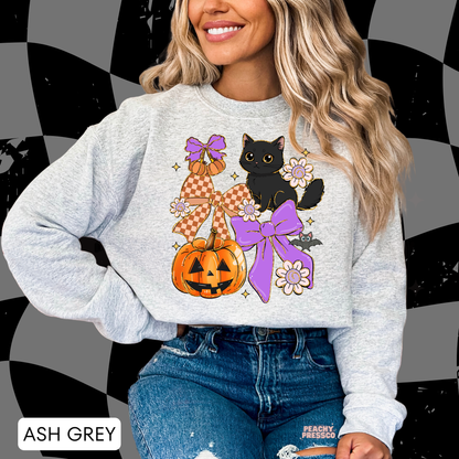 Woman wearing an ash grey crewneck featuring a cute black cat, jack-o'-lantern pumpkin, purple bows, flowers and bat Halloween graphic.