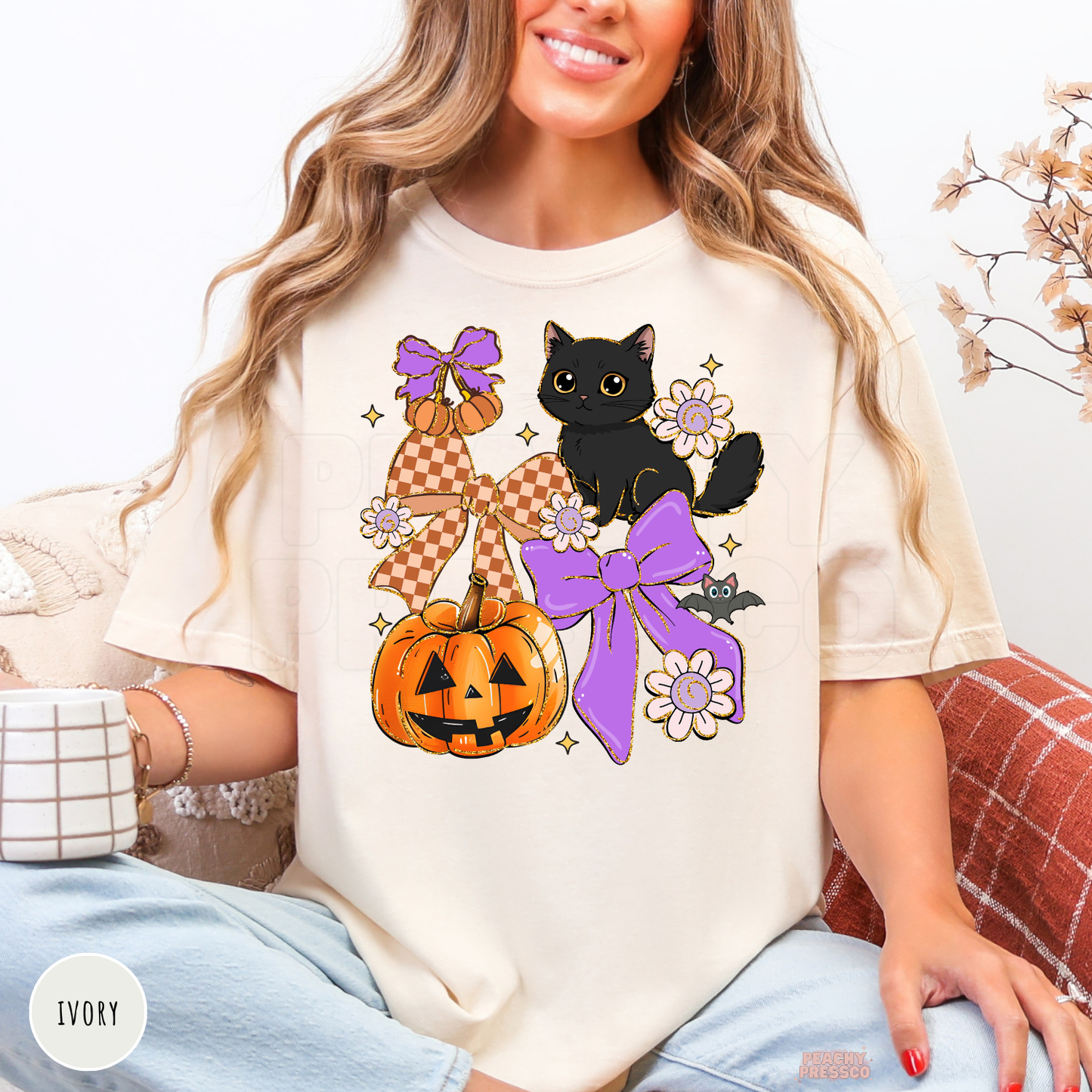 Woman wearing an ivory Comfort Colors t-shirt featuring a black cat, pumpkin, purple bows, floral accents and bat Halloween artwork.