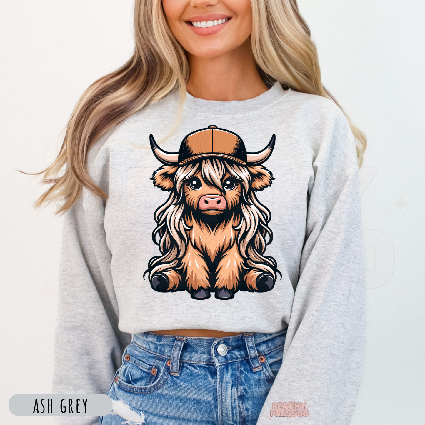 Cute Highland cow wearing a trucker hat printed on an ash grey crewneck sweatshirt with a western country-inspired graphic.