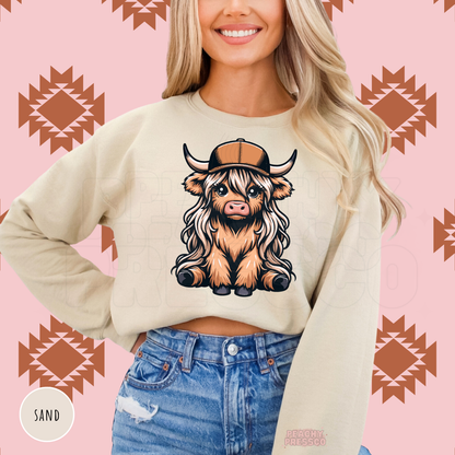 Cute Highland cow wearing a brown trucker hat displayed on a sand-colored western crewneck sweatshirt.
