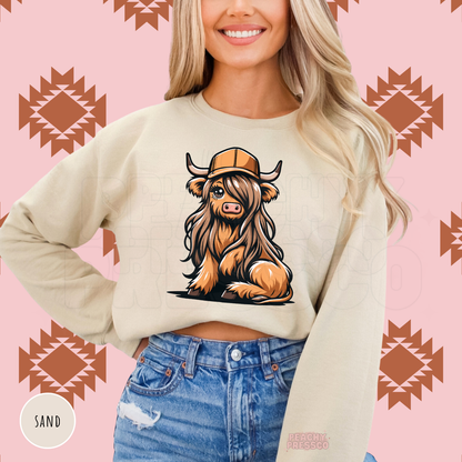 Cute Highland cow with long hair and a tan trucker hat on a sand colored western graphic sweatshirt.