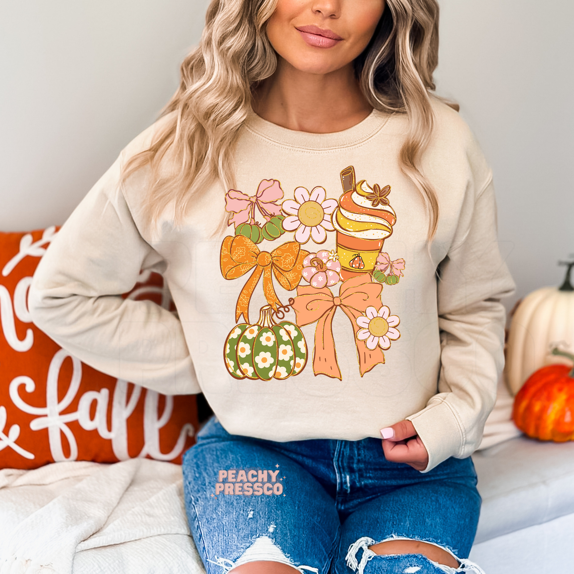 Woman wearing a sand crewneck sweatshirt featuring a cute coquette fall graphic with bows, pumpkin spice drink, daisy flowers, and decorative pumpkins in soft autumn colors.