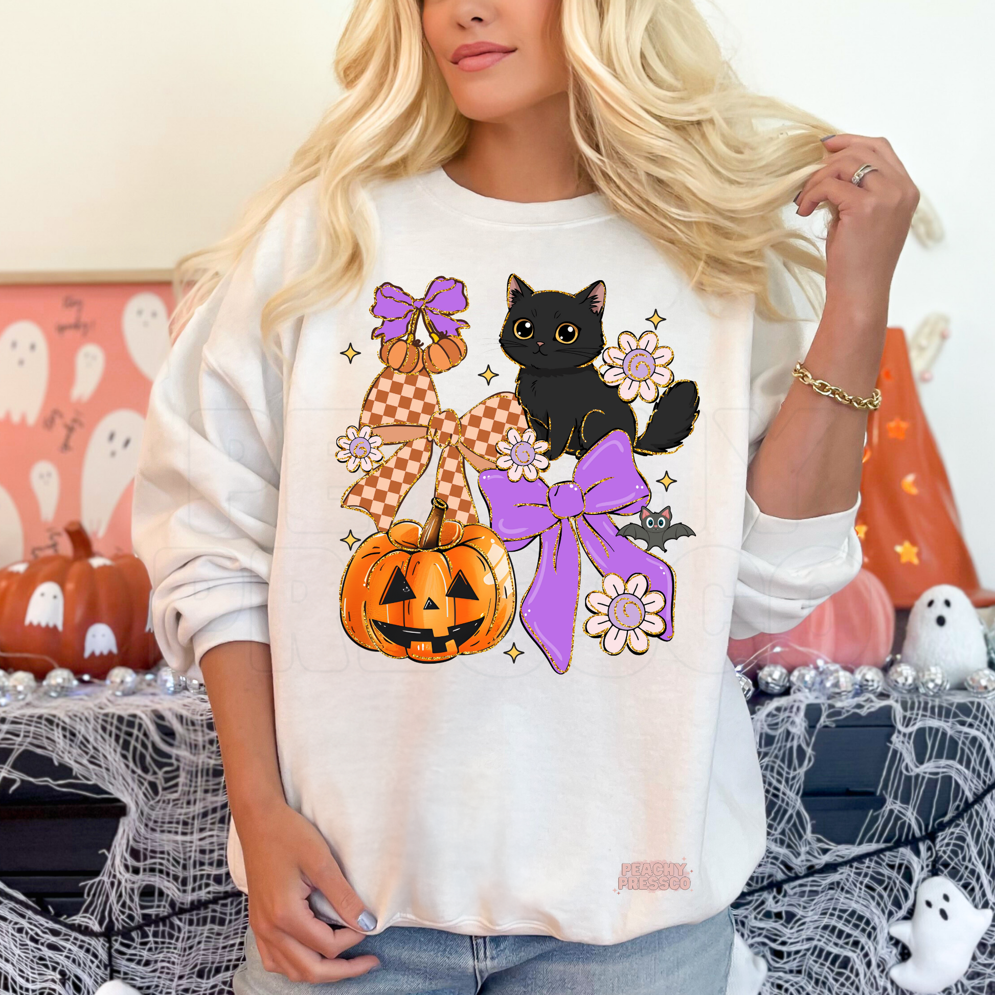 Woman wearing a white Halloween crewneck with a black kitten, pumpkin, purple bows, flowers and cute bat graphic.