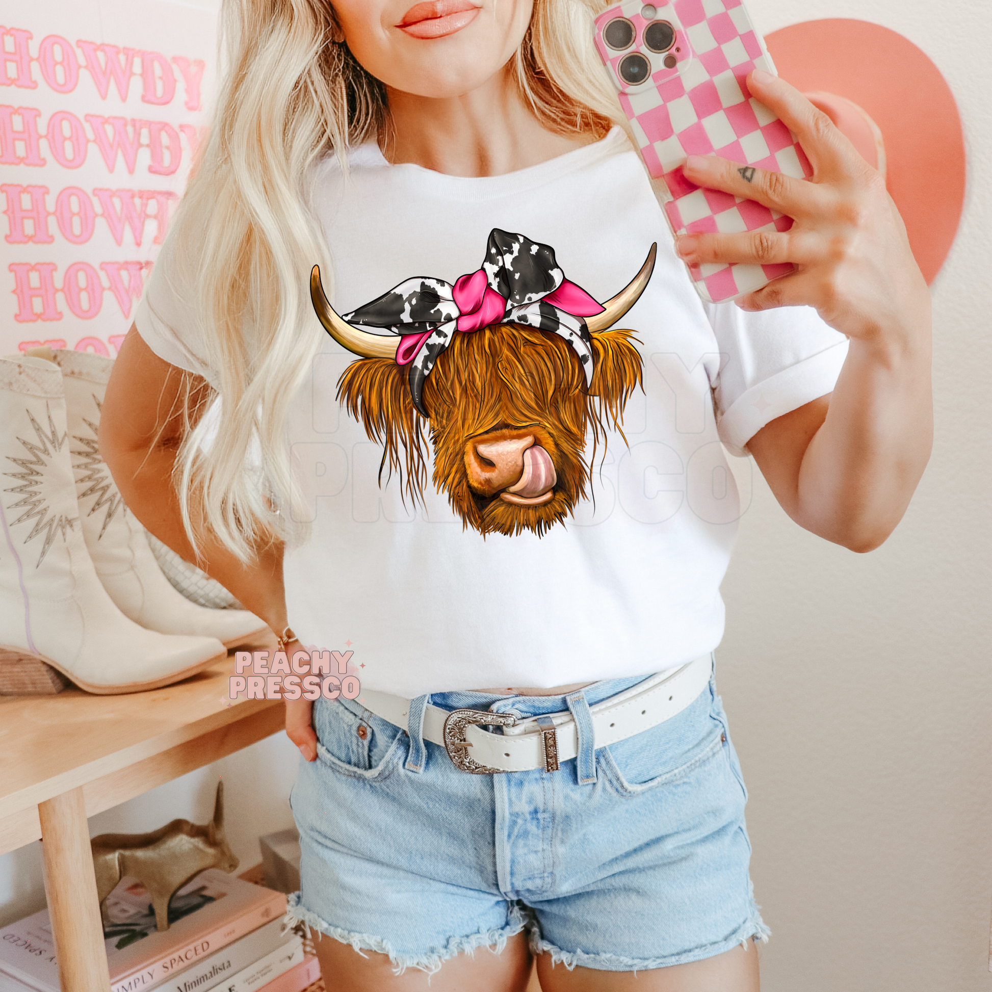 White graphic tee featuring a Highland cow with a black and white cow print bandana, bright pink bow, and playful tongue.