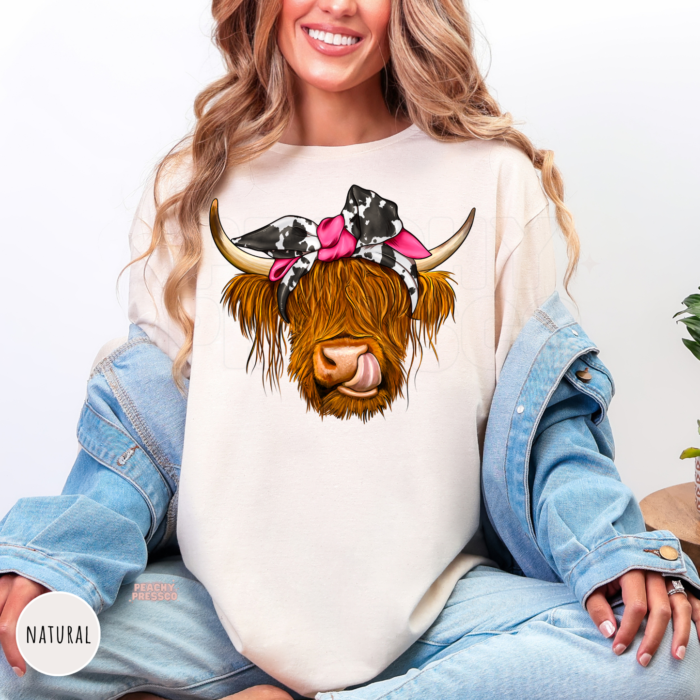 Natural color shirt featuring a funny Highland cow with a cow print bandana, pink bow, shaggy hair, and playful tongue western graphic.