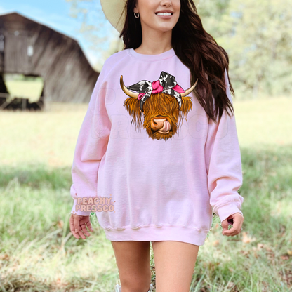 Light pink sweatshirt with a cute Highland cow graphic featuring a cow print bandana, pink bow, and western country aesthetic.