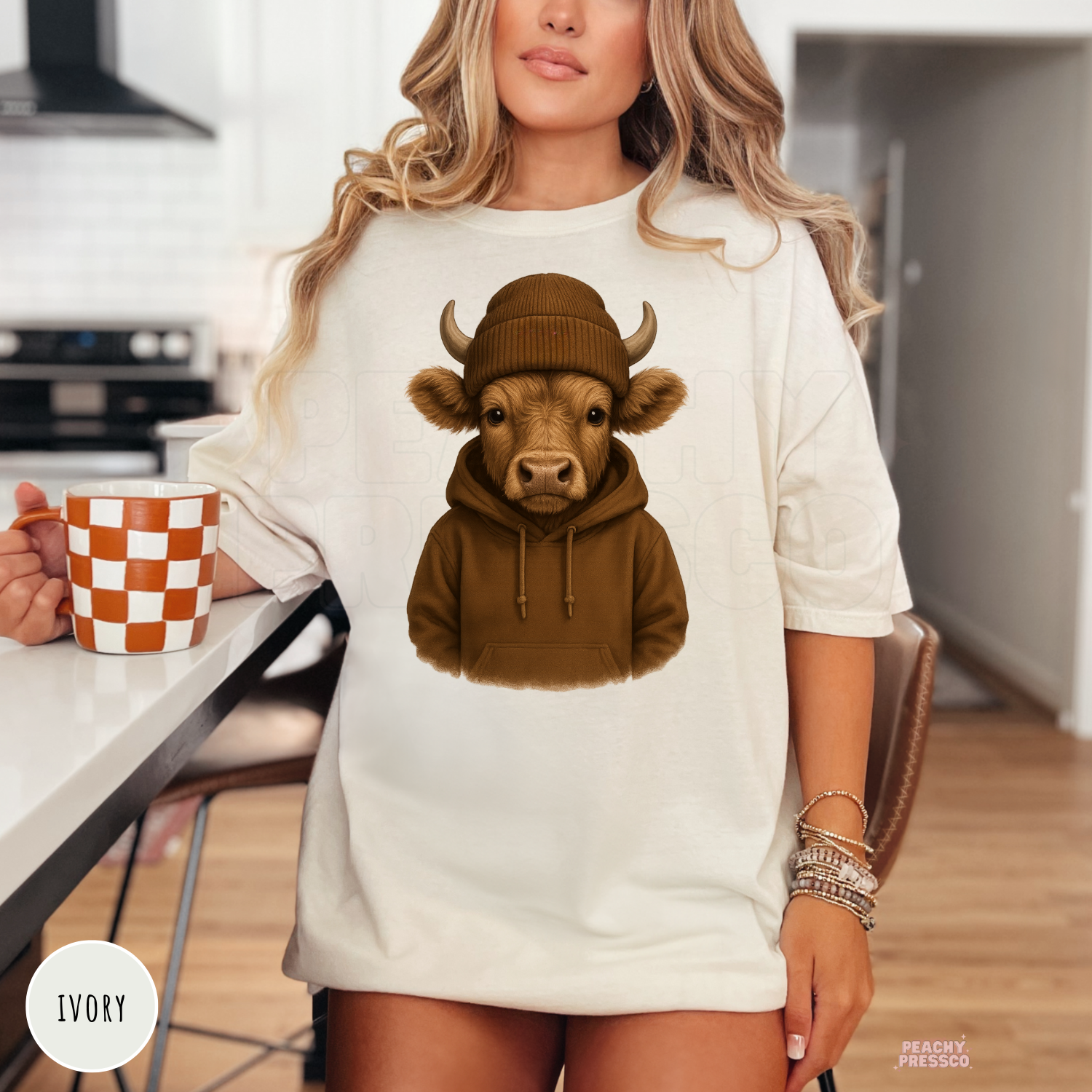 Woman wearing an ivory t-shirt featuring a cute Highland cow wearing a brown hoodie and knit beanie, cozy western farm animal graphic.