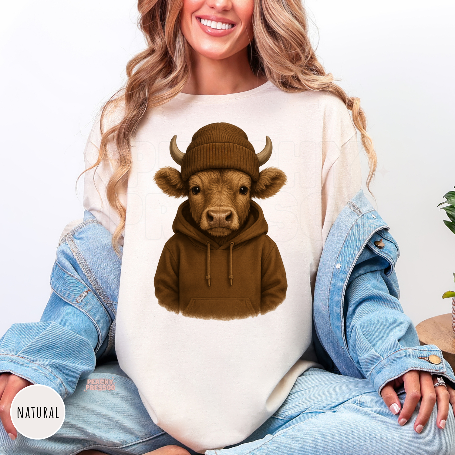 Woman wearing a natural shirt featuring a cute Highland cow wearing a cozy brown hoodie and knit beanie, rustic western design.