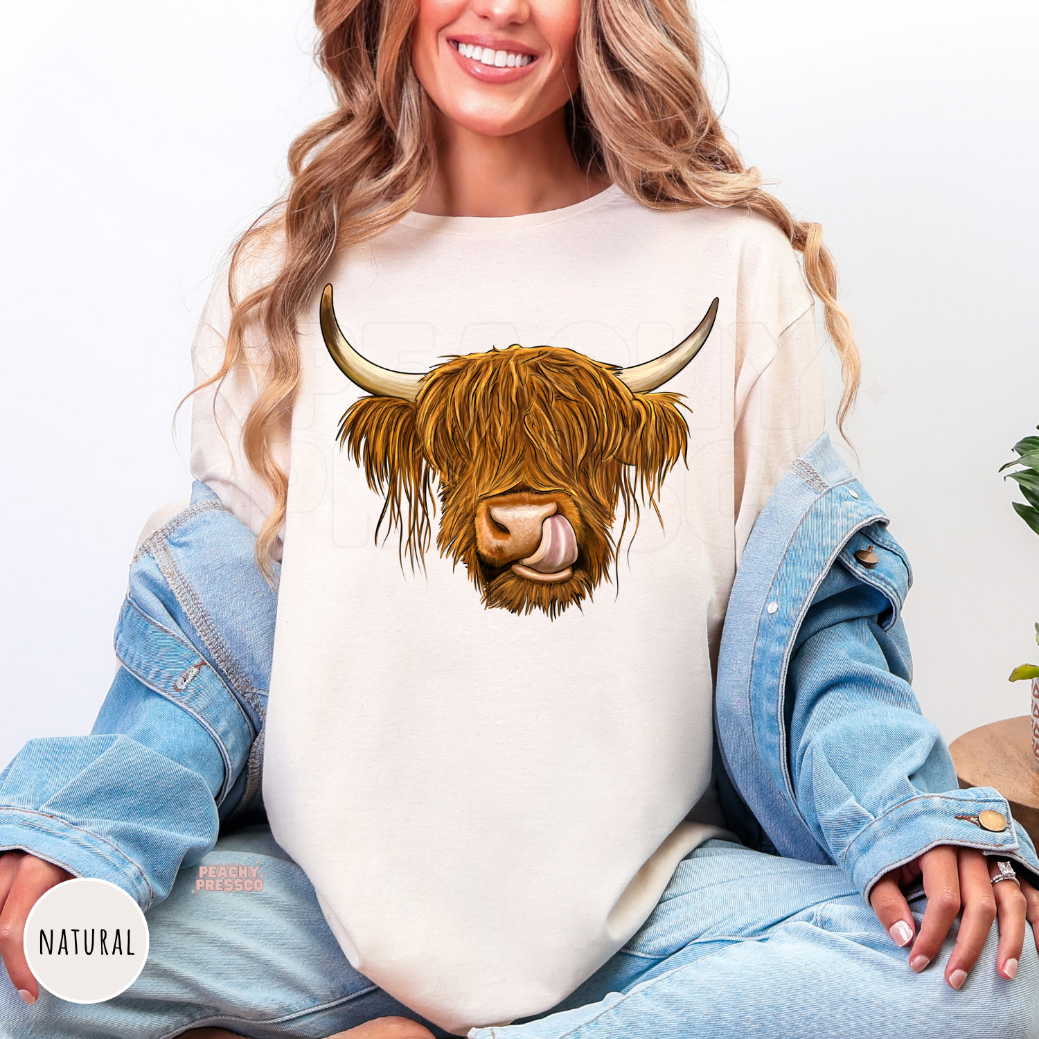 Woman wearing a natural-colored shirt featuring a Highland cow graphic with long shaggy hair and rustic farmhouse style.