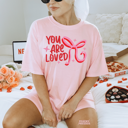 Soft pink oversized t-shirt with a cheerful "You Are Loved" Valentine's quote and coquette bow graphic.
