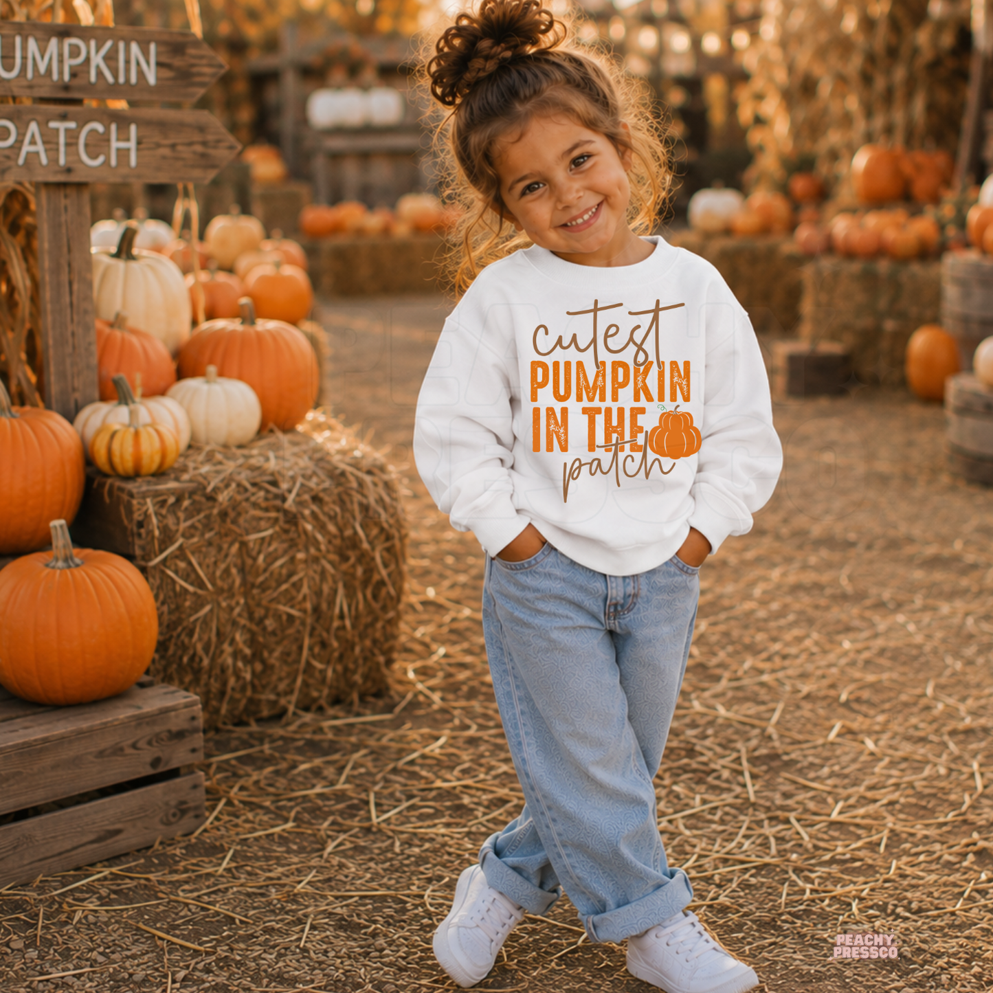 Toddler girl wearing a white crewneck sweatshirt featuring a "Cutest Pumpkin in the Patch" graphic while standing at a pumpkin patch.
