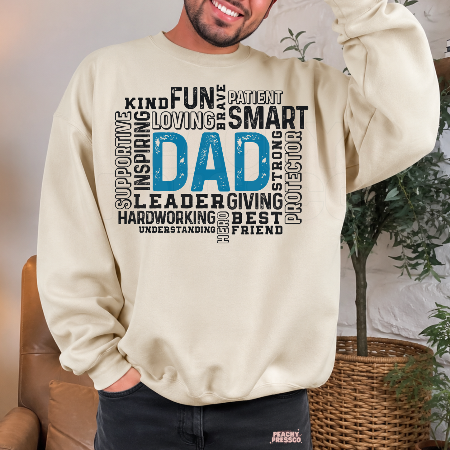 Sand crewneck sweatshirt featuring a bold blue "Dad" word art design surrounded by meaningful words including kind, smart, supportive, loving, hardworking, leader, protector, and best friend. A thoughtful Father's Day gift for dads.