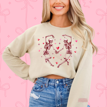 Sand crewneck sweatshirt with a whimsical dancing skeleton couple graphic accented by pink hearts and sparkling details.