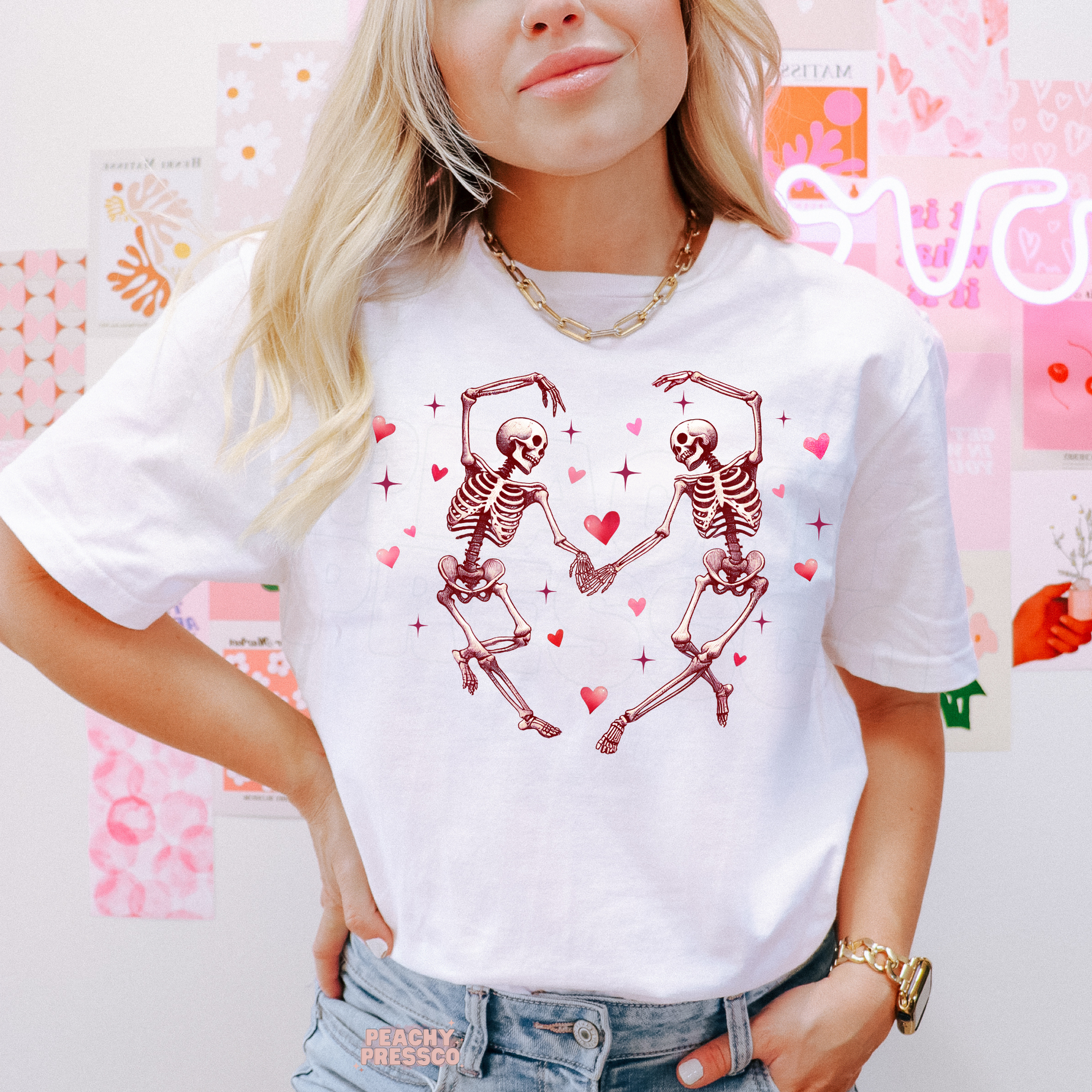 White graphic t-shirt featuring two dancing skeletons holding hands with pink hearts in a playful gothic love design.