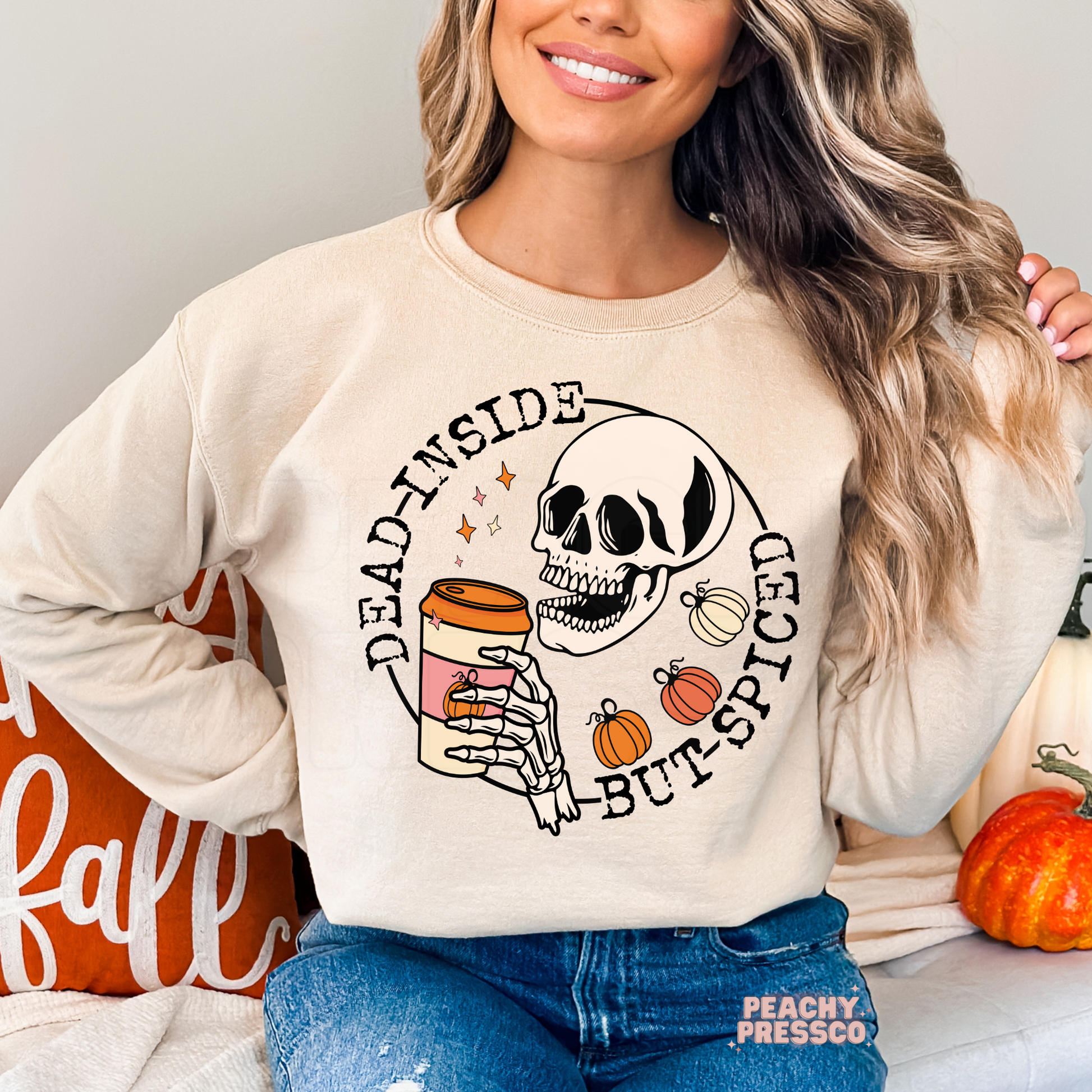 Woman wearing a sand crewneck sweatshirt featuring a skeleton drinking pumpkin spice coffee with colorful fall pumpkins.