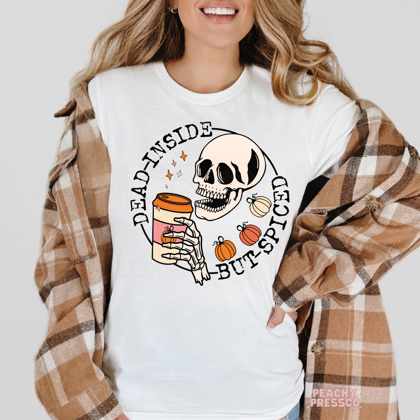 Woman wearing a white graphic t-shirt featuring a skeleton holding pumpkin spice coffee with the phrase "Dead Inside But Spiced."