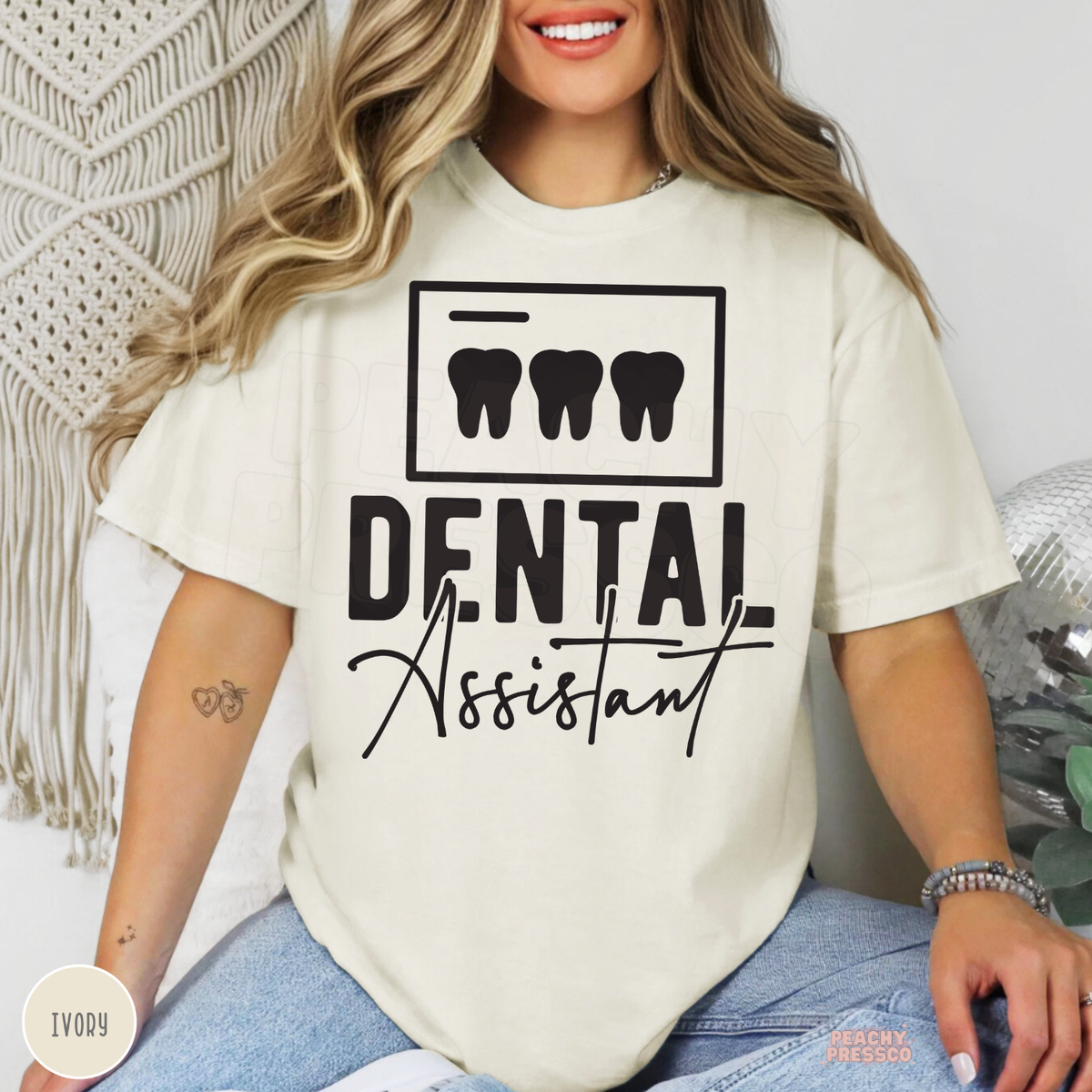 Woman modeling an ivory Dental Assistant T-shirt with black tooth icons and dental assistant lettering, ideal for dental team gifts.