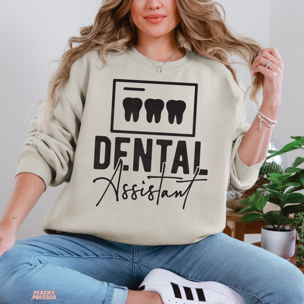 Woman wearing a sand colored Dental Assistant crewneck sweatshirt featuring three tooth icons and bold black dental assistant lettering.