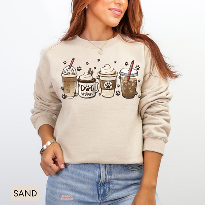 Woman modelling a sand-coloured Dog Mom coffee sweatshirt with illustrated coffee drinks, a dog mom mug and paw print details.