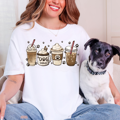 Woman wearing a white Dog Mom coffee graphic T-shirt featuring four illustrated coffee cups and paw prints while sitting beside her dog.