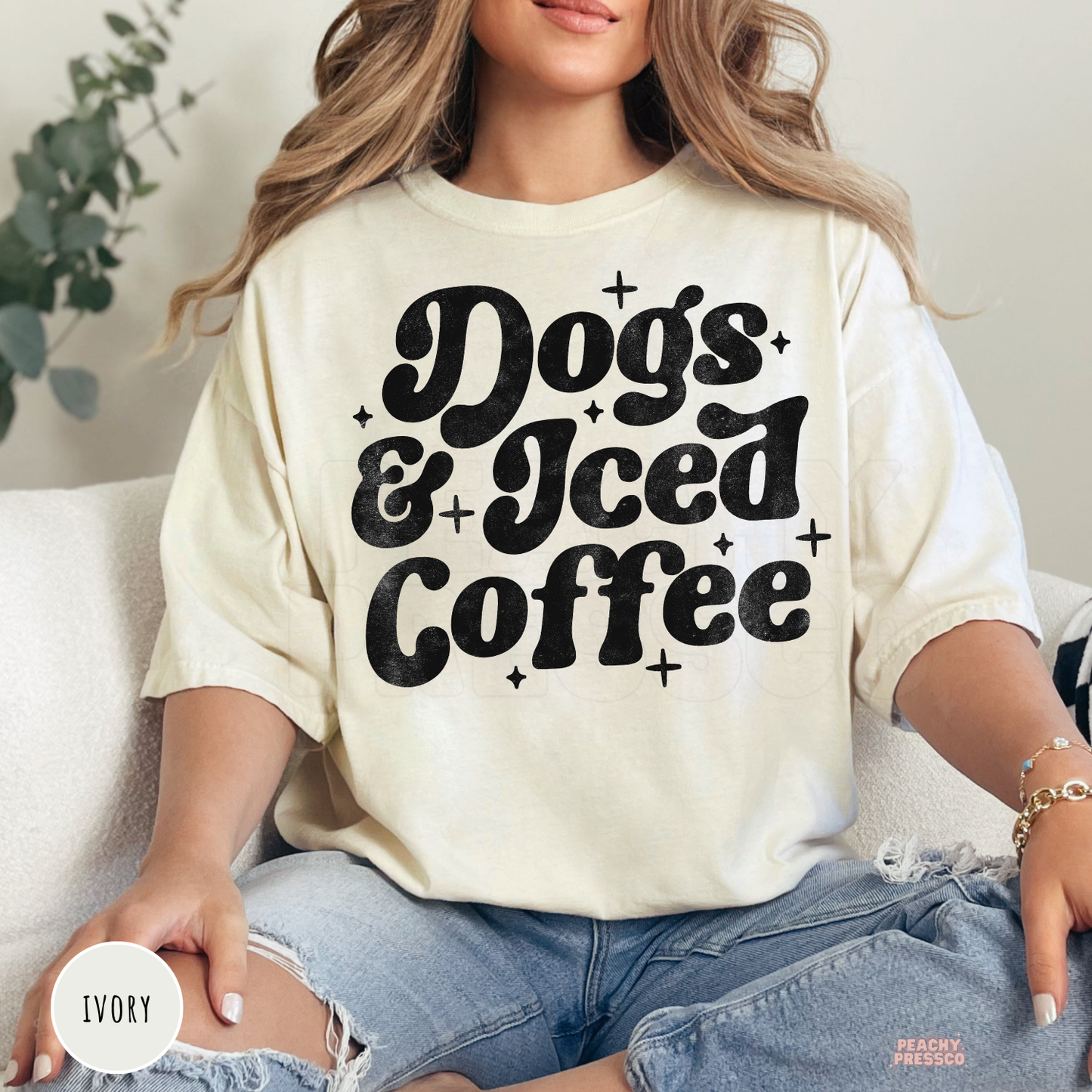 Woman wearing an ivory oversized t-shirt featuring a retro "Dogs & Iced Coffee" graphic with distressed lettering.
