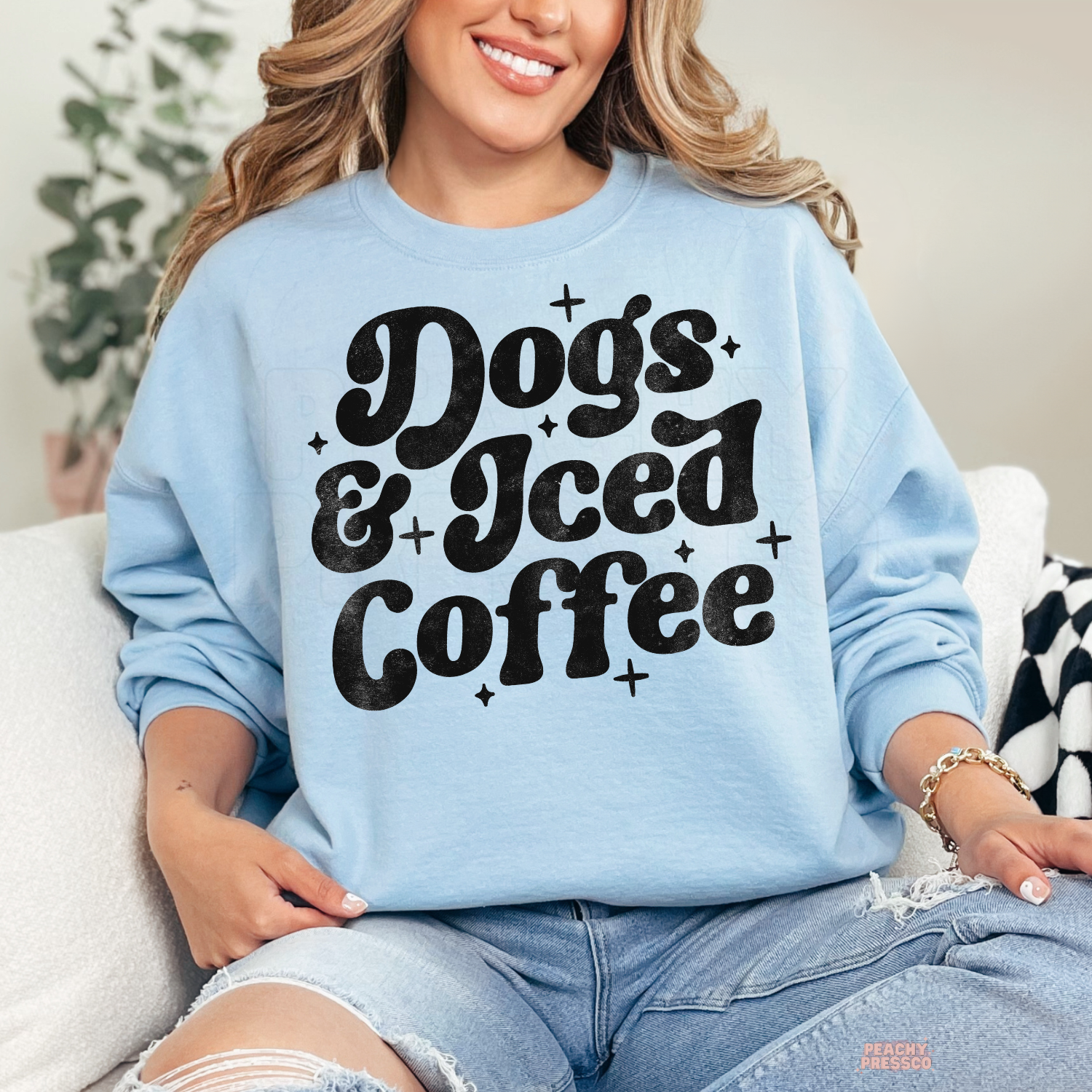 Woman wearing a light blue crewneck sweatshirt featuring a bold retro "Dogs & Iced Coffee" graphic.
