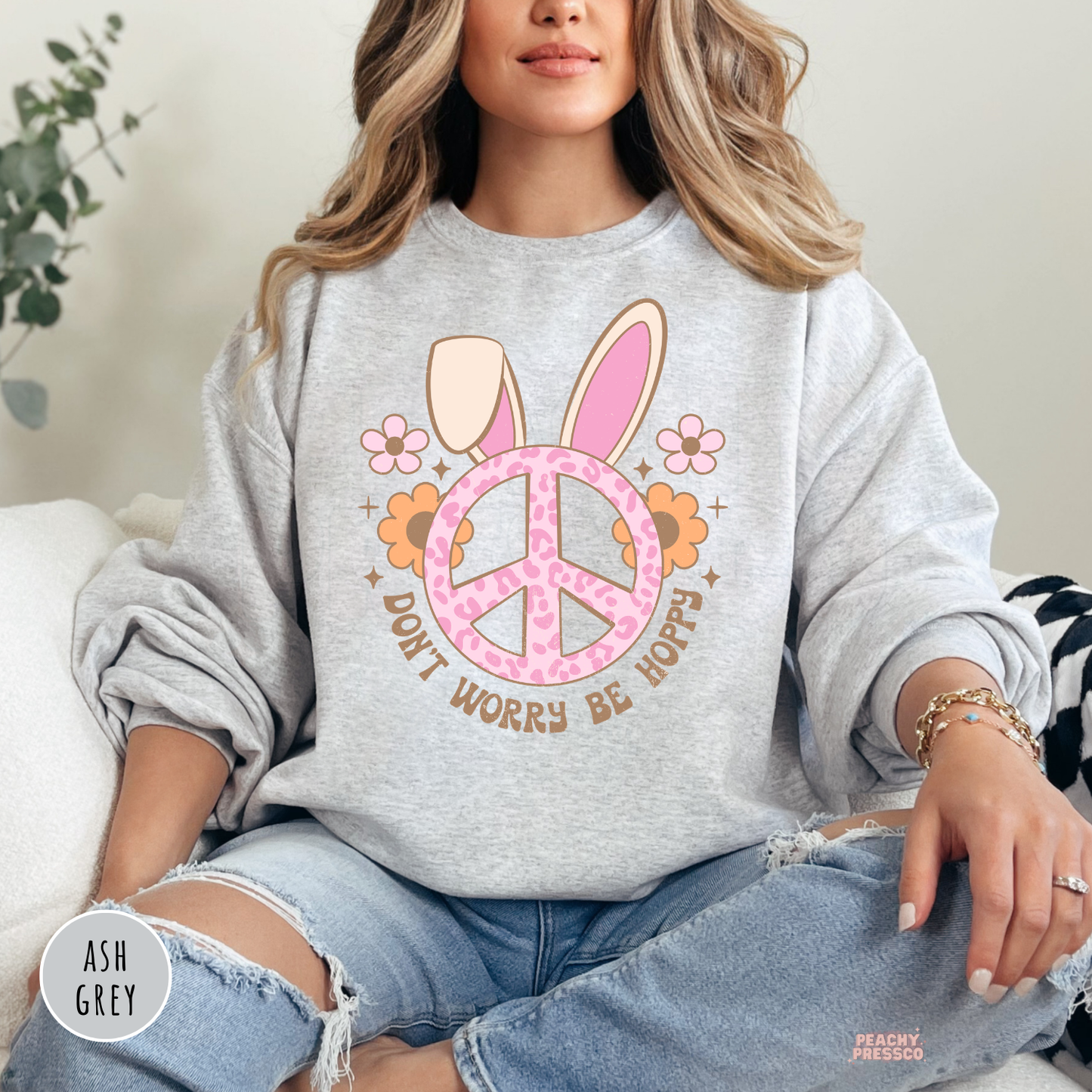 Woman wearing an ash grey crewneck sweatshirt featuring a pink leopard print peace sign with bunny ears and the phrase "Don't Worry Be Hoppy" for Easter.