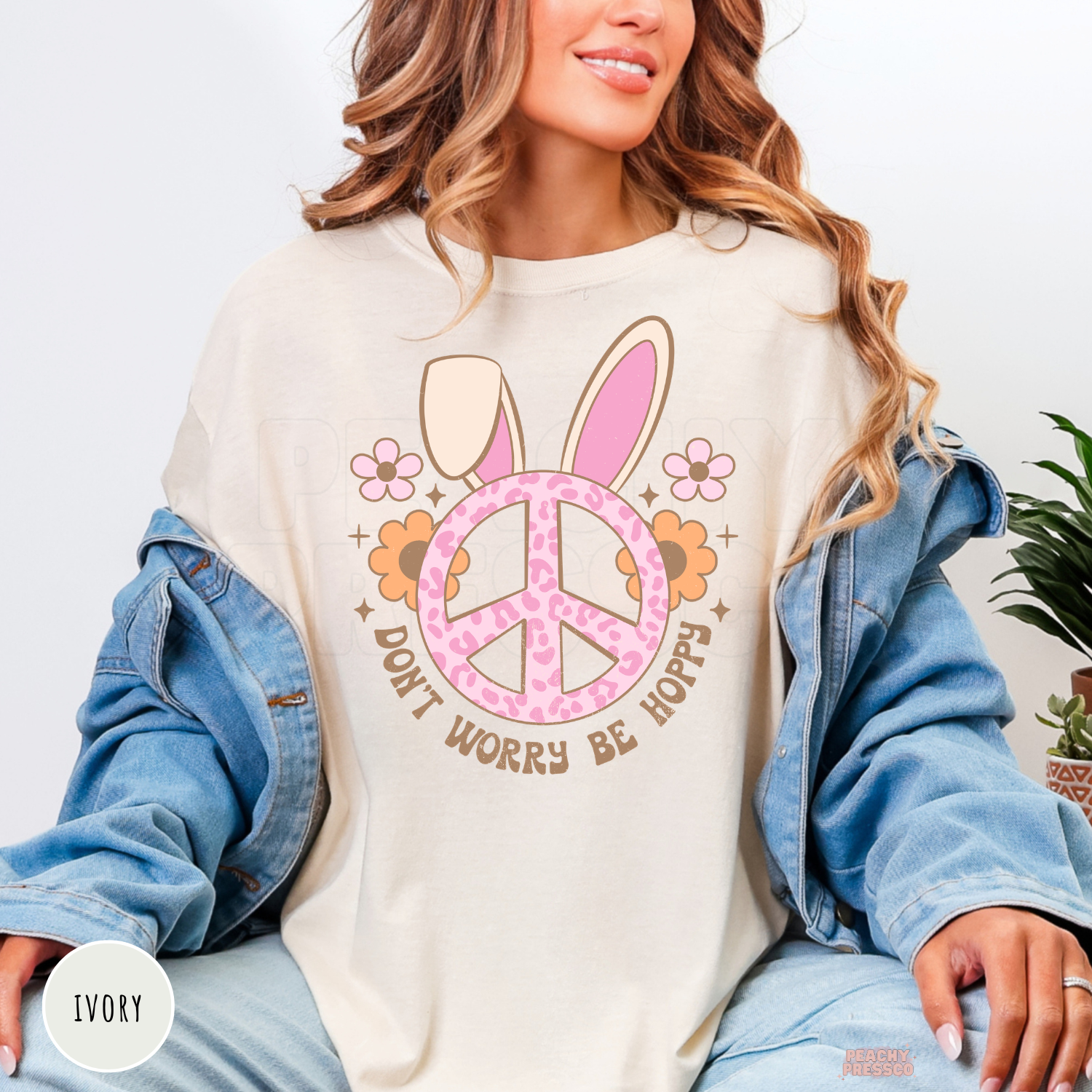Woman wearing an ivory Comfort Colors Easter t-shirt featuring a pink peace sign with bunny ears, flowers, and "Don't Worry Be Hoppy."