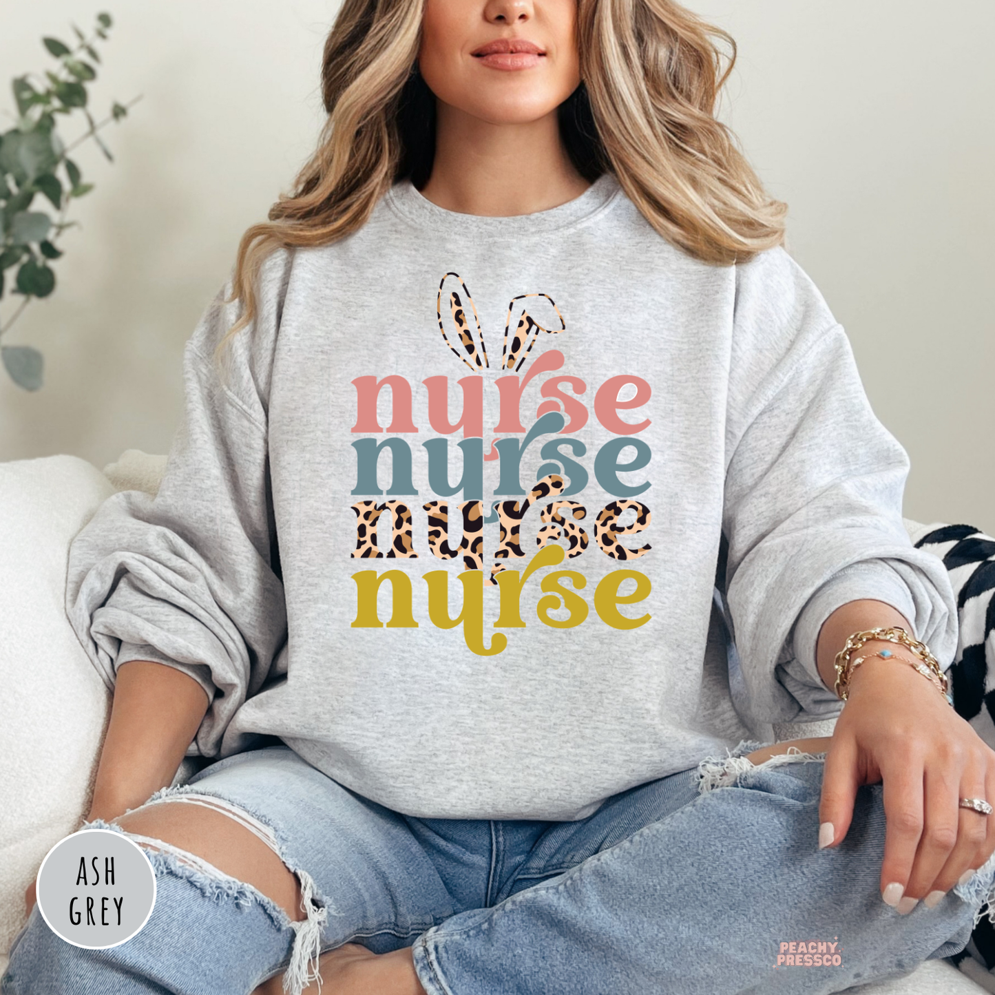 Woman wearing an ash grey crewneck sweatshirt featuring a retro "Nurse" Easter design with bunny ears, leopard print lettering, and pastel spring colors.