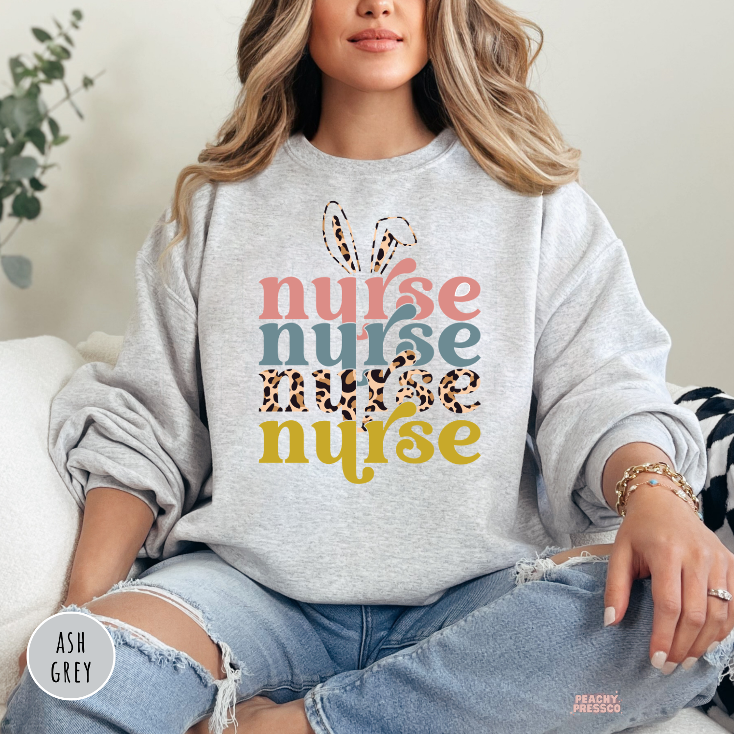 Woman wearing an ash grey crewneck sweatshirt featuring a retro "Nurse" Easter design with bunny ears, leopard print lettering, and pastel spring colors.
