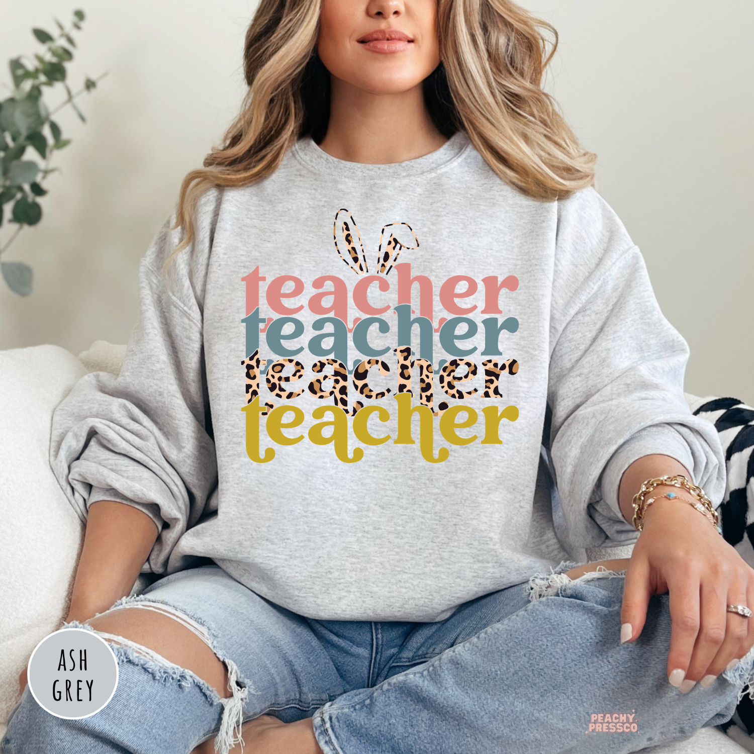 Woman wearing an ash grey crewneck sweatshirt featuring a retro Easter Teacher design with bunny ears, leopard print lettering, and pastel spring colors.